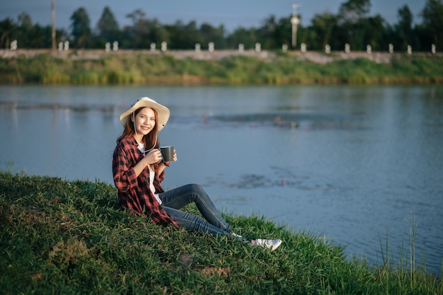 Jeune femme asiatique en chapeau de randonnée assise près du lac et tenant une tasse de café, Sourire se détendre pendant le voyage de camping