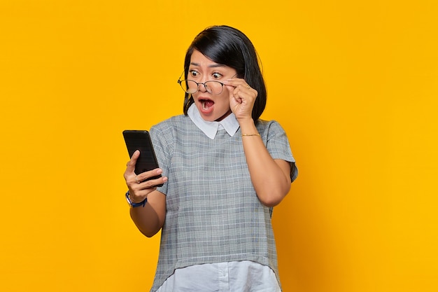 Jeune femme asiatique choquée utilisant un téléphone portable et prenant des lunettes isolées sur fond jaune