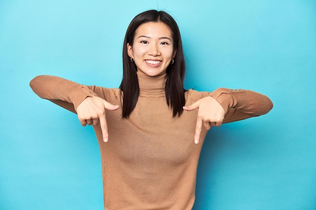 Jeune femme asiatique en col roulé marron pointe vers le bas avec les doigts sentiment positif