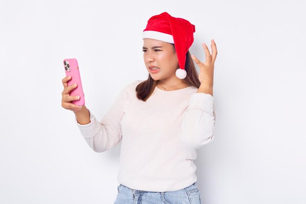 Jeune femme asiatique en colère dans un chapeau de Noël regardant smartphone obtenir de bonnes nouvelles isolé sur fond blanc isolé sur fond blanc célébration vacances de Noël et concept du nouvel an