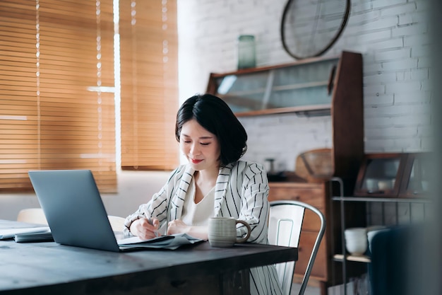 Jeune femme asiatique concentrée travaillant à domicile en utilisant un ordinateur portable et en manipulant des documents dans le salon à la maison Mode de vie flexible