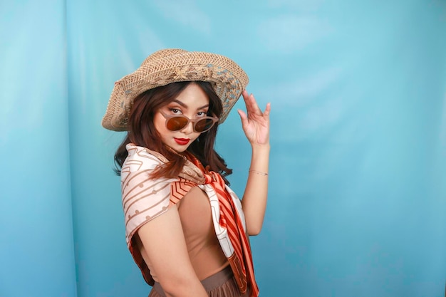 Une jeune femme asiatique confiante et heureuse regarde et tient ses lunettes de soleil isolées par un fond bleu