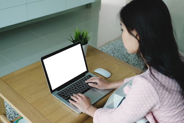 Jeune femme asiatique créative assise à son bureau à l'aide d'un ordinateur portable avec une maquette d'écran blanc dans le salon confortable d'une maison moderne.