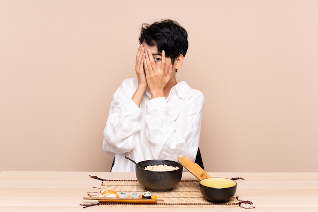 Jeune femme asiatique dans une table avec bol de nouilles et sushi couvrant les yeux et regardant à travers les doigts