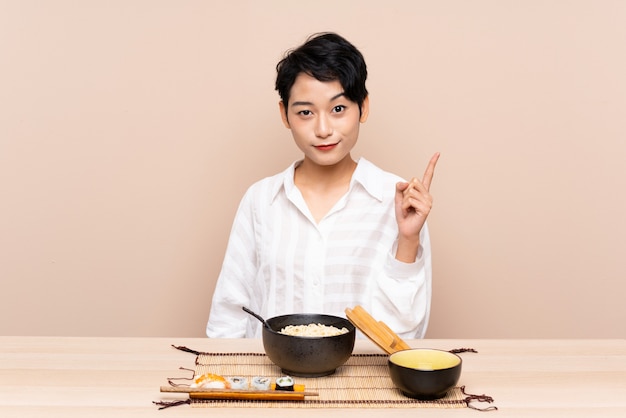 Jeune femme asiatique dans une table avec un bol de nouilles et de sushis pointant avec l'index une excellente idée