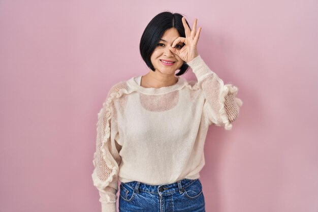 Jeune femme asiatique debout sur fond rose faisant un geste correct avec la main souriant oeil regardant à travers les doigts avec un visage heureux