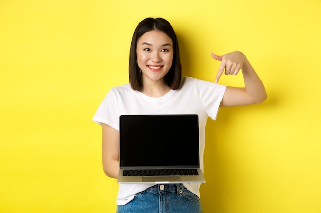 Jeune femme asiatique démontre une offre en ligne, montrant un écran d'ordinateur portable et souriant, debout sur fond jaune