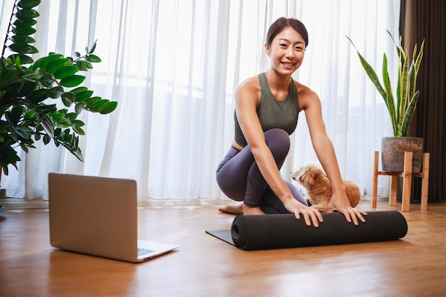 Jeune femme asiatique déroulant un tapis de yoga noir pour la formation en ligne de yoga avec son chien à la maison