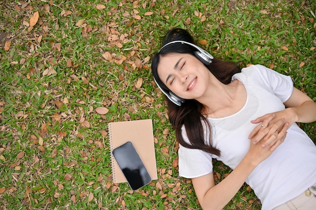 Jeune femme asiatique détendue et effrayante écoutant de la musique souriant les yeux fermés et allongée sur l'herbe