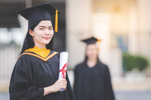 Une jeune femme asiatique diplômée d'université en robe de graduation et en mortier détient un certificat de diplôme devant le bâtiment de l'université après avoir participé à la rentrée universitaire