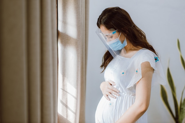 Jeune femme asiatique enceinte portant un écran facial et un masque médical debout près de la fenêtre à la maison pour l'auto-quarantaine de la propagation des maladies Covid 19