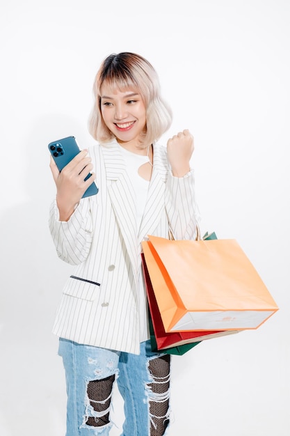 Jeune femme asiatique énergique tenant un smartphone vierge à la recherche d'un magasin de détail avec des sacs à provisions colorés sur fond blanc Concept futuriste de magasinage en ligne