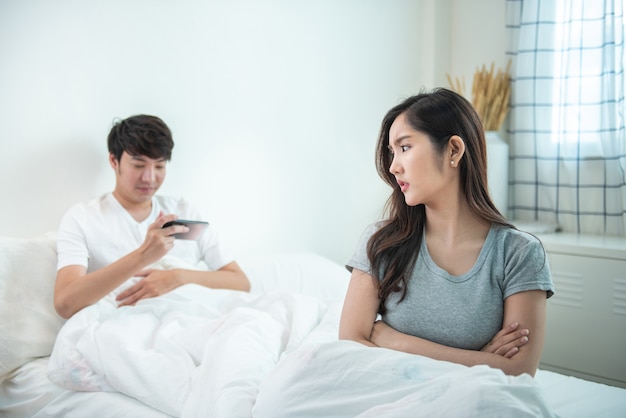 La jeune femme asiatique est bouleversée par le fait que l'homme utilise tout le temps son téléphone sans lui prêter attention. L'homme tient le téléphone prêter attention au téléphone mobile intelligent tout en étant assis sur le lit en ignorant sa petite amie.