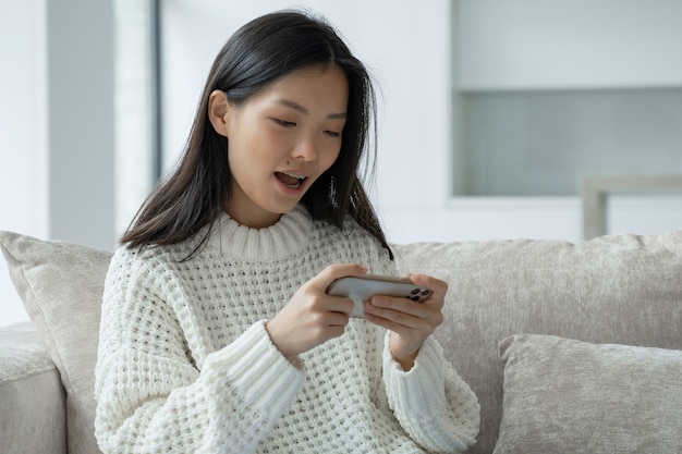 Une jeune femme asiatique excitée est assise sur le canapé du salon et joue à un jeu mobile sur un
