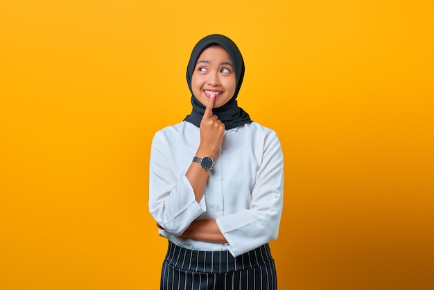 Jeune femme asiatique excitée touchant les lèvres et ayant des idées sur fond jaune