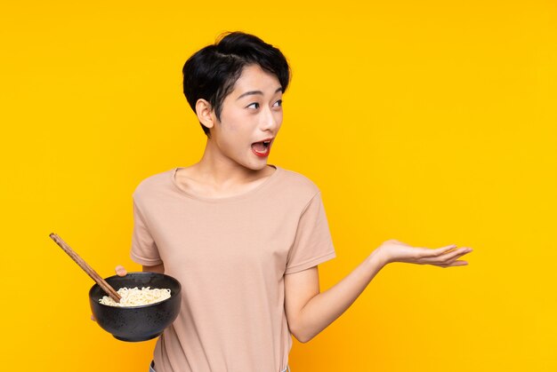 Jeune femme asiatique avec une expression faciale surprise tout en tenant un bol de nouilles avec des baguettes