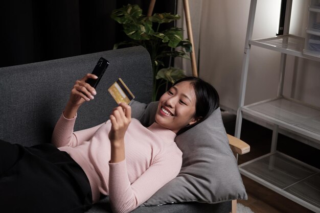 Une jeune femme asiatique fait des achats en ligne avec sa carte de crédit alors qu'elle est assise sur le canapé à la maison.