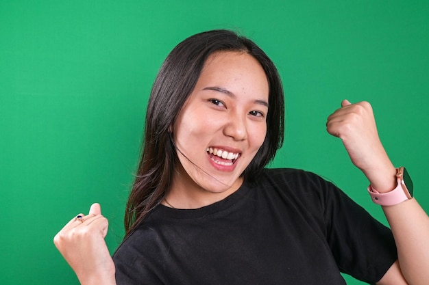 Une jeune femme asiatique fait le geste du gagnant et serre les poings en disant oui sur un fond vert.