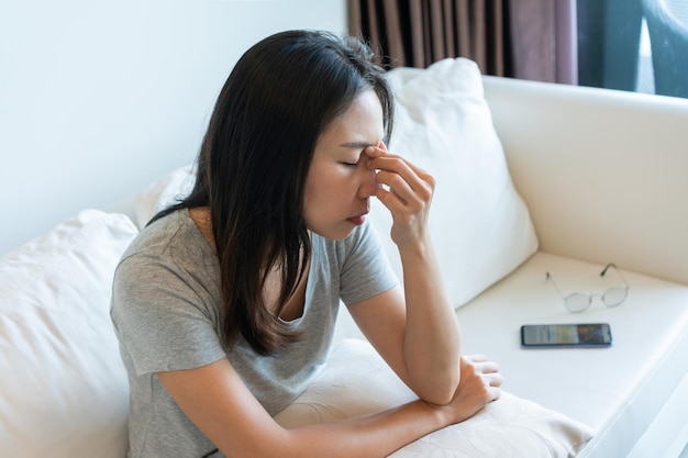 Jeune femme asiatique fatiguée qui enlève ses lunettes et ressent de la fatigue oculaire lors de l'utilisation d'un téléphone portable à la maison Fille souffrant de fatigue du syndrome de l'œil sec après une longue utilisation du téléphone intelligent