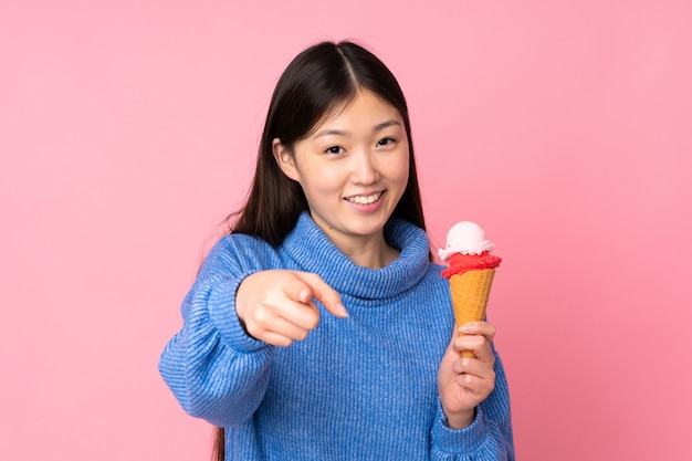 Jeune femme asiatique avec une glace au cornet isolé sur le mur rose pointe le doigt vers vous avec une expression confiante