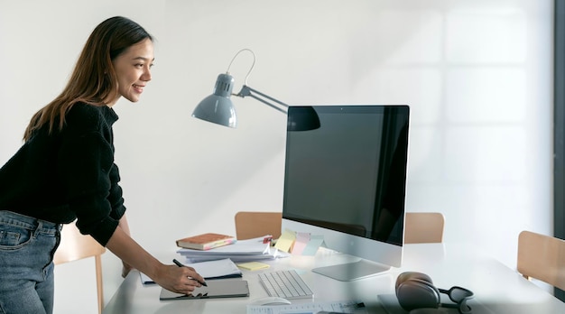 Jeune femme asiatique heureuse debout à son bureau à l'aide d'une tablette numérique et d'un ordinateur de bureau