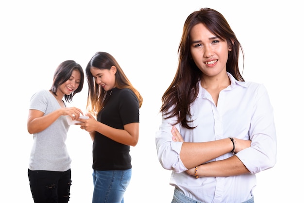 Jeune femme asiatique heureuse souriant avec les bras croisés avec deux amis heureux à l'arrière à l'aide de téléphone mobile ensemble