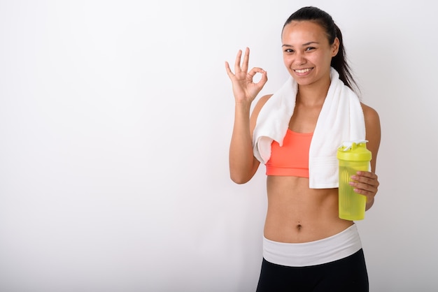 jeune femme asiatique heureuse souriant tout en donnant signe ok et tenant une bouteille d'eau avec une serviette autour du cou prêt pour la salle de sport contre l'espace blanc.