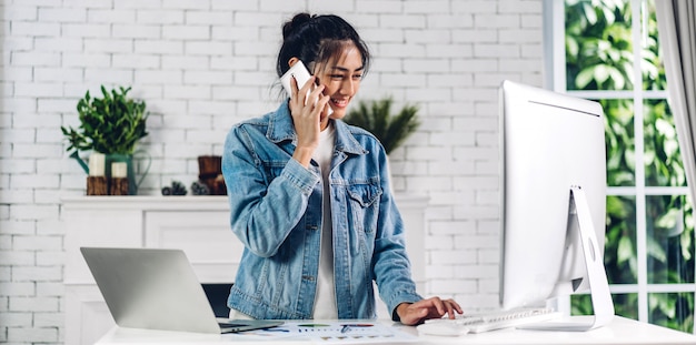 Jeune femme asiatique heureuse souriante relaxante à l'aide d'un ordinateur de bureau et d'une réunion de vidéoconférence en ligne chat à la maison.une fille créative utilise un smartphone et tape sur le clavier.travail à partir du concept de la maison