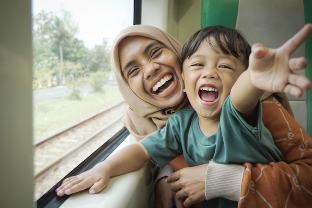 Jeune femme asiatique en hijab et son fils riant joyeusement et regardant par la fenêtre à l'intérieur du train