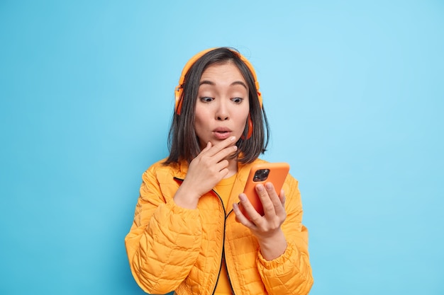 Une jeune femme asiatique impressionnée, concentrée sur un smartphone, écoute de la musique via un casque et réagit à quelque chose d'incroyable vêtue d'une élégante veste orange isolée sur un mur bleu. Technologie moderne