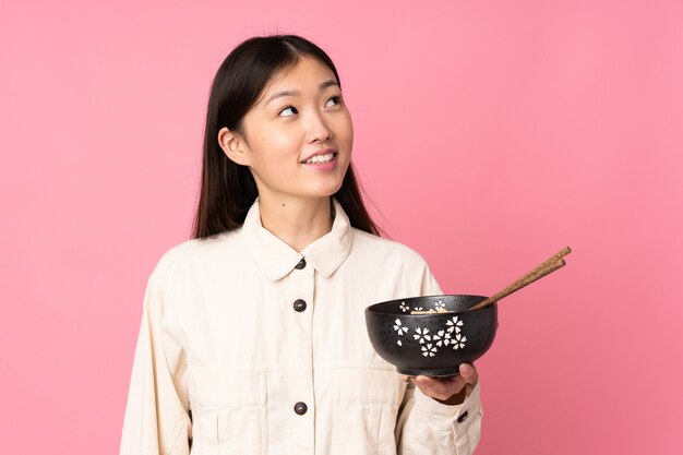 Jeune femme asiatique isolée sur fond rose en levant tout en souriant tout en tenant un bol de nouilles avec des baguettes