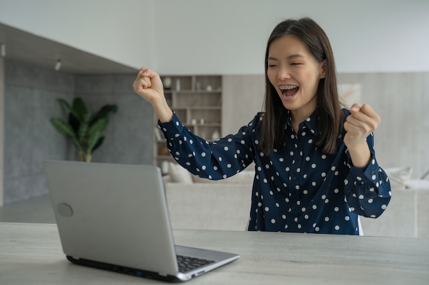 Une jeune femme asiatique joyeuse et enthousiaste est assise à une table avec un ordinateur portable, lève les mains et célèbre