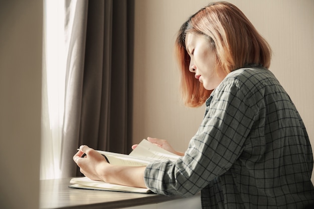 Jeune femme asiatique lisant un livre près de la fenêtre.