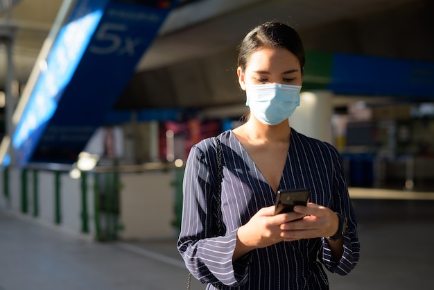 Jeune femme asiatique avec masque à l'aide de téléphone tout en marchant de la gare