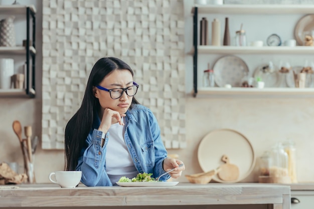 Jeune femme asiatique mécontente en lunettes et chemise en jean assise dans la cuisine à la maison à la table