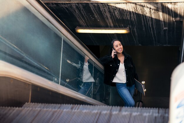 Une jeune femme asiatique monte un escalator à la station de métro avec une planche à roulettes à la main tout en parlant au téléphone