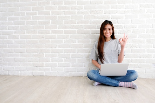 Jeune femme asiatique montrant le signe de la main ok et souriant tout en travaillant avec un ordinateur portable dans la salle blanche