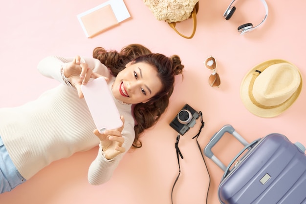 Jeune femme asiatique montrant son smartphone en position allongée sur un sol rose avec un entourage de bagages de voyage