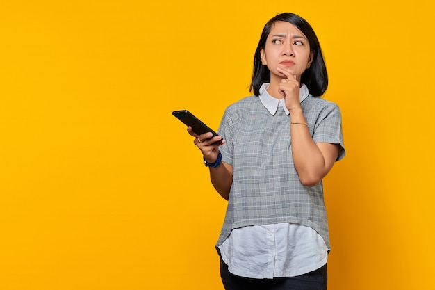 Jeune femme asiatique pensive utilisant un téléphone portable avec les doigts sur le menton ang regardant de côté