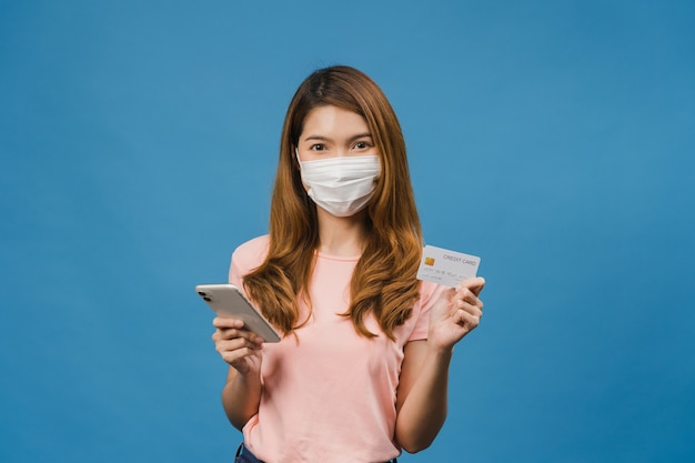 Jeune femme asiatique portant un masque médical utilisant un téléphone et une carte bancaire de crédit avec une expression positive, sourit largement, vêtue de vêtements décontractés et se tient isolée sur un mur bleu