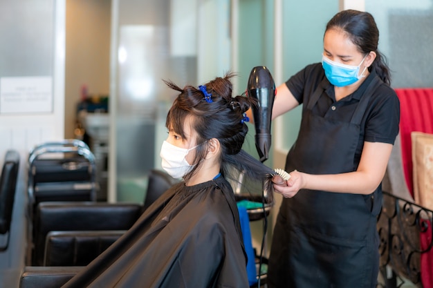 Jeune femme asiatique portant des masques pour se protéger de Covid-19 pendant la coiffure à l'aide d'un sèche-cheveux et d'un peigne pour cheveux noirs dans un salon de beauté.