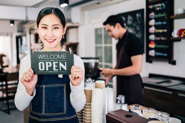 Jeune femme asiatique propriétaire d'un café barista tenant le panneau d'annonce "BIENVENUE", "OUVERT" avec un barista masculin flou en arrière-plan. Affaires avec un partenaire. Travail d'équipe d'entrepreneur réussi.
