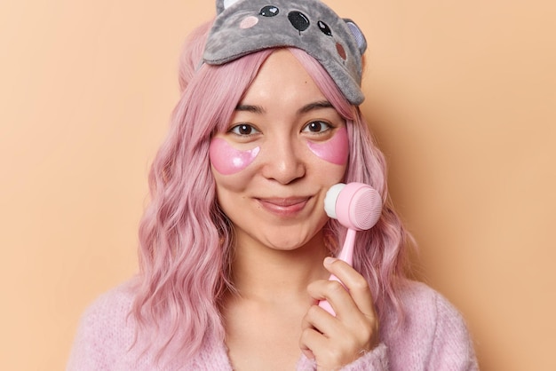 Une jeune femme asiatique ravie aux cheveux roses utilise un massage du visage applique des patchs d'hydrogel sous les yeux pour éliminer les ridules porte un masque de sommeil isolé sur fond beige. Concept de soins du visage et de beauté