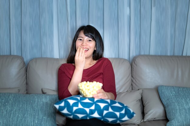 Jeune femme asiatique, regarder la télévision suspense film ou des nouvelles à la recherche de manger du pop-corn heureux tard dans la nuit à la maison salon canapé.