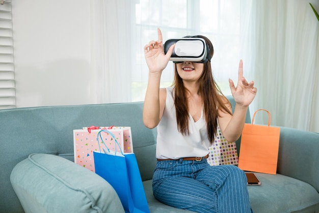 Jeune femme asiatique avec des sacs de shopping portant des casques de réalité virtuelle modernes ayant de l'expérience dans le shopping en ligne dans le salon à la maison Femme heureuse avec un casque VR sur le canapé