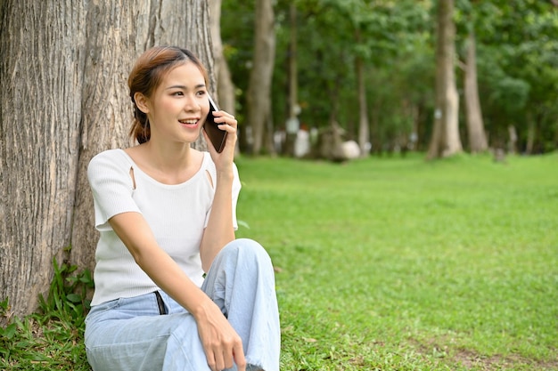 Une jeune femme asiatique se détend assise sous l'arbre et au téléphone en appelant quelqu'un