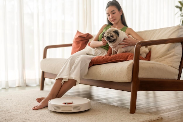 Une jeune femme asiatique se détend avec un chien pug dans le salon pendant que l'aspirateur robotique travaille