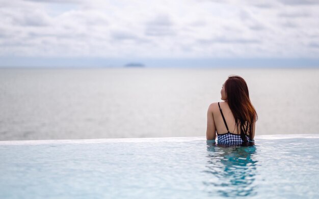 Une jeune femme asiatique se relaxant dans une piscine à débordement en regardant une belle vue sur la mer