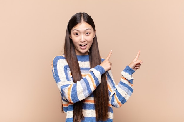 Jeune femme asiatique se sentant joyeuse et surprise, souriant avec une expression choquée et pointant vers le côté