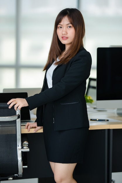 Jeune femme asiatique séduisante en affaires noires debout avec confiance dans un bureau moderne avec un arrière-plan flou de fenêtres. Concept de style de vie de bureau moderne.